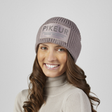 Pikeur Beanie Selection 285800 Variant Ash Mauve. A smiling woman wearing a ribbed knit beanie in a muted taupe-gray color with a rectangular front patch reading 'PIKEUR'. She is also wearing a light beige ribbed turtleneck sweater against a plain light gray studio background.