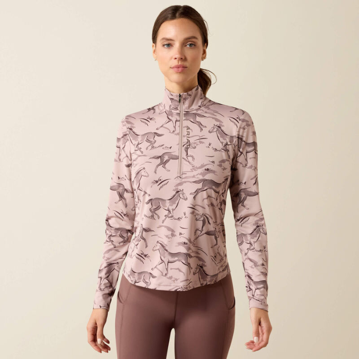 Ariat Ladies Lunaveil 1/2 Zip Long Sleeve Base Layer Variant Sphinx Meadow Frolic Print. A woman modeling a long-sleeve quarter-zip athletic top with an allover horse print in dusty pink and taupe, paired with mauve leggings, against a solid light beige background.