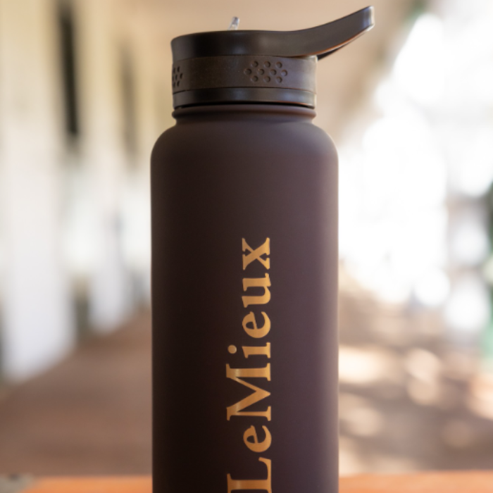 LeMieux AW26 Drinks Bottle Varient Cocoa. A dark brown insulated water bottle with a black flip-top lid and vertical gold 'LeMieux' branding, shown against a softly blurred indoor background.