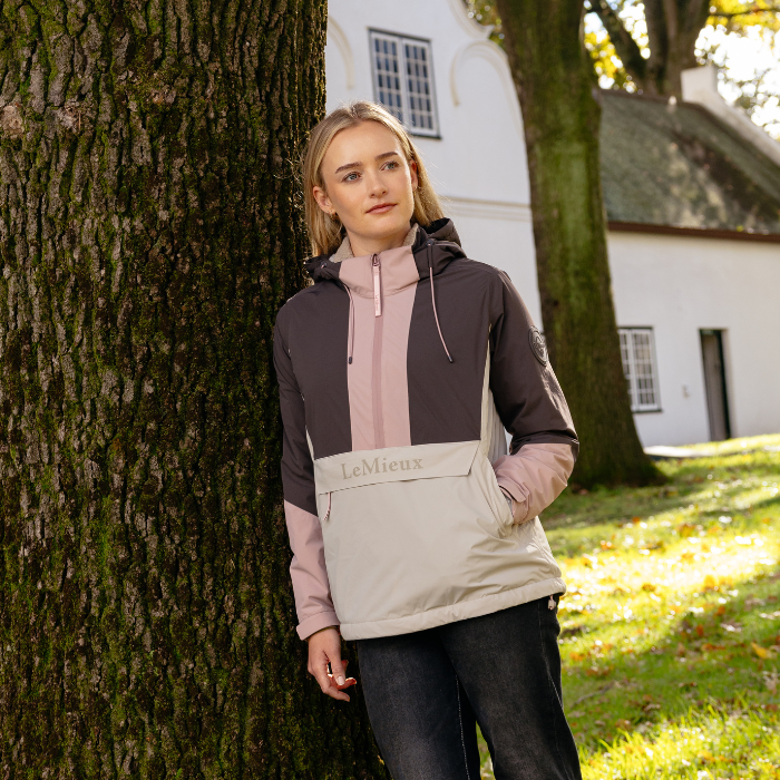 LeMieux AW26 Phoebe Half Zip Waterproof Anorak Varient Wild Rose. A woman stands outdoors leaning against a large tree trunk, wearing a color-blocked hooded jacket. The jacket features a dark gray upper section, a dusty pink central panel and sleeves, and a light beige lower panel with the brand name 'LeMieux' printed on it.