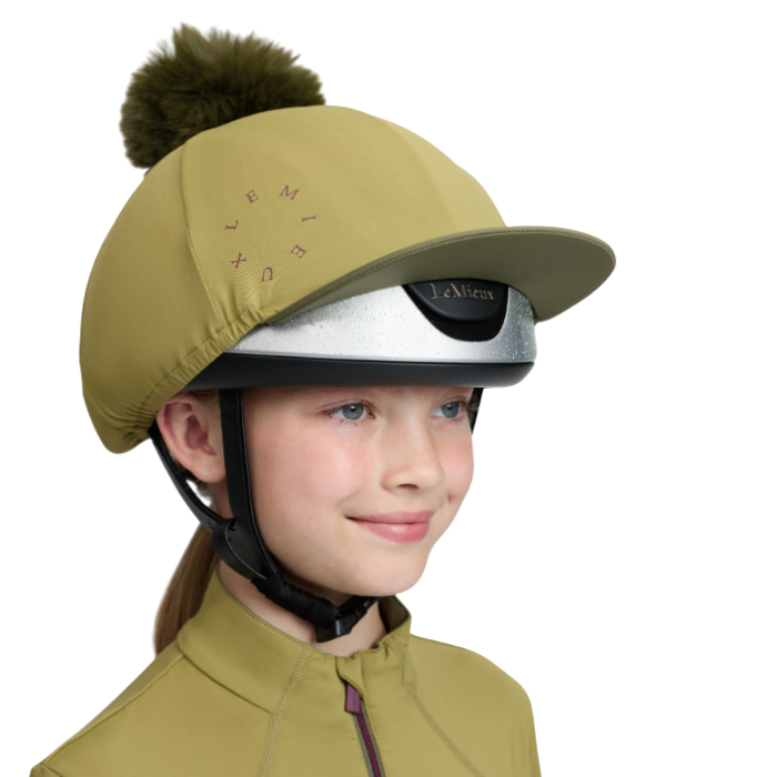 LeMieux AW26 Classique Pom Hat Silk Variant Colour Gooseberry.
A child wearing a gooseberry green equestrian riding helmet cover with a matching pom-pom, a silver riding helmet, and a matching riding jacket, shown against a plain white background.