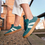 Teal sneakers with white stripes and brown soles worn by a person sitting on a railing.