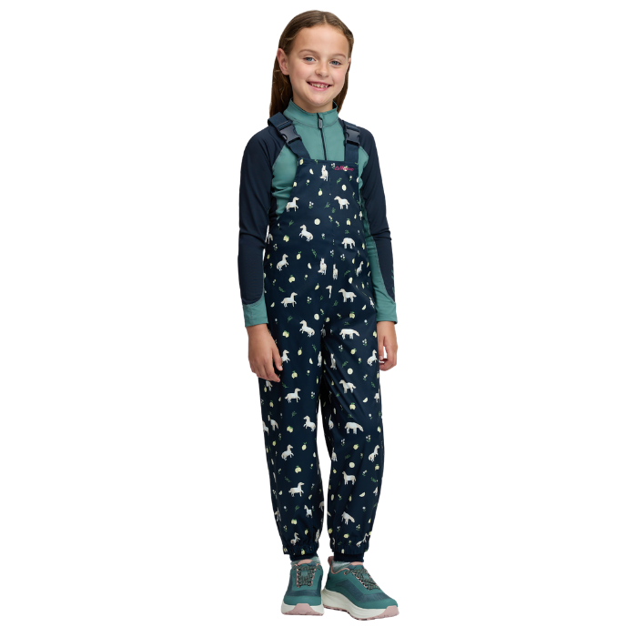 LeMieux AW26 Mini Pippin Splash Dungarees Varient Navy. A young girl stands against a plain white background wearing a dark navy overall with a white horse print, layered over a teal and navy long-sleeve shirt, and teal sneakers.