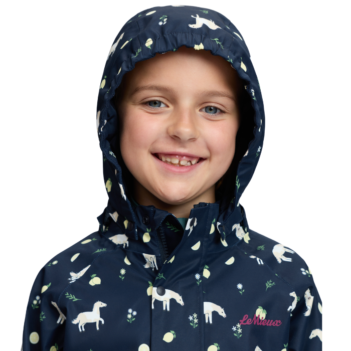 LeMieux AW26 Mini Pippin Splash Jacket Varient Navy. A smiling child wearing a navy hooded rain jacket with a white and yellow animal and floral print, photographed against a plain white background.