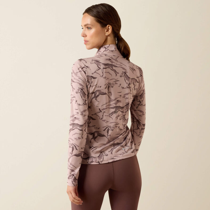 Ariat Ladies Lunaveil 1/2 Zip Long Sleeve Base Layer Variant Sphinx Meadow Frolic Print. A woman shown from the back wearing a long-sleeve athletic top with an allover horse pattern in dusty mauve and brown, paired with brown leggings, against a plain beige studio background.