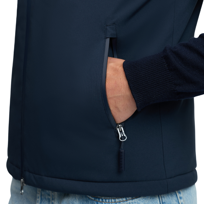 LeMieux AW26 Mens Midweight Insulated Gilet Varient Navy. Close-up of a person wearing a dark navy zippered jacket, with a hand in a side pocket and a ribbed navy sleeve visible, against a plain light background.