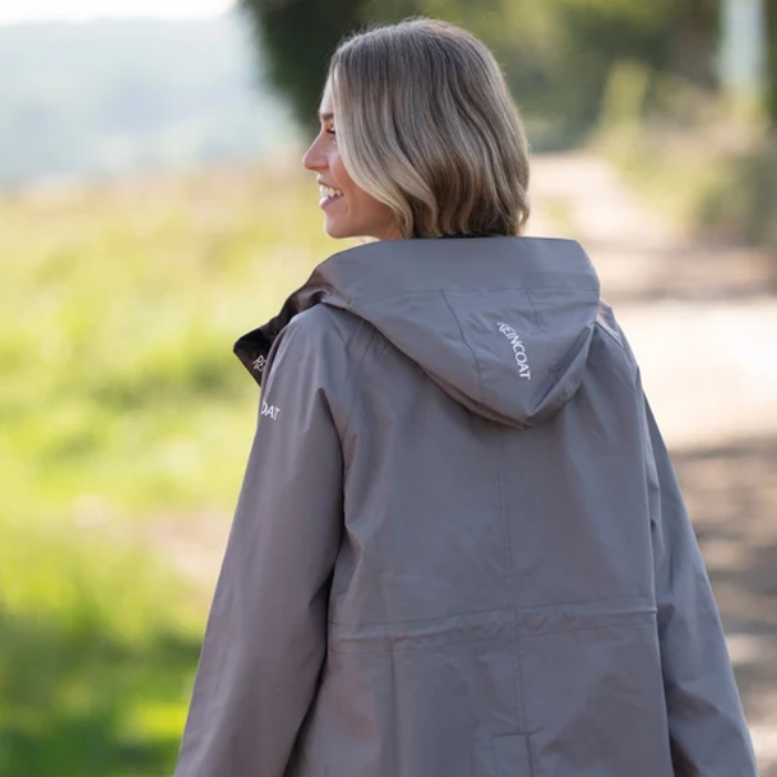 Reincoat Parka Lite Varient Dune. A woman wearing a gray hooded rain jacket, photographed from the back and side in an outdoor setting with a blurred natural background.