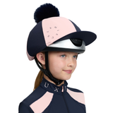 LeMieux AW26 Harlequin Hat Silk Variant Colour Navy/Petal.
A child wearing a navy and light pink equestrian helmet with a matching pom-pom and a matching navy and light pink riding jacket, shown against a plain white background.