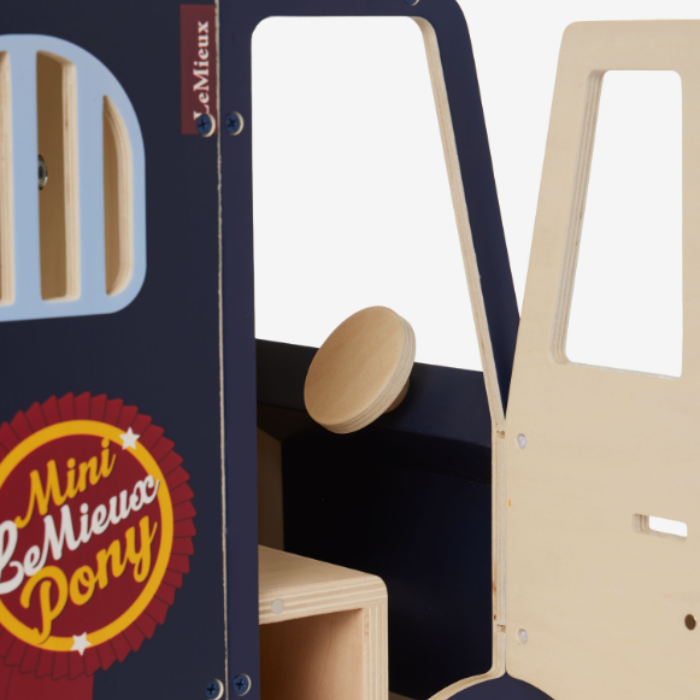 LeMieux AW26 Toy Pony Lorry Wooden Varient Brown. Close-up of a children's wooden play set featuring a dark navy blue panel with a round window, a beige wooden door, and a wooden seat. A red and yellow 'Mini LeMieux Pony' sticker is visible on the front panel.