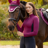LeMieux AW26 Classique Base Layer Variant Colour Blackberry.
A woman in a fitted long-sleeve riding top and dark breeches stands beside a brown horse wearing a matching magenta fly veil and saddle pad, with a blurred outdoor background.