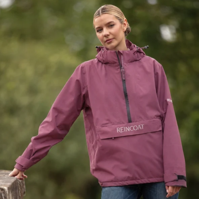Reincoat Smock Lite Varient Bloom. Woman wearing a Berry coloured jacket with a black zip and kangaroo style pocket with "REINCOAT" branding on a blurred green background, leaning against a wall. 