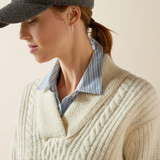 Woman wearing a cream sweater with a striped shirt underneath and a dark cap.
