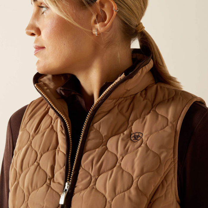 Close-up of a person wearing a brown quilted vest with a brand logo on a neutral background