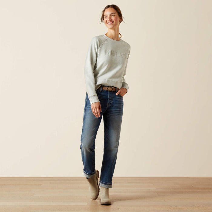 Woman wearing a light grey sweatshirt and blue jeans standing against a plain background