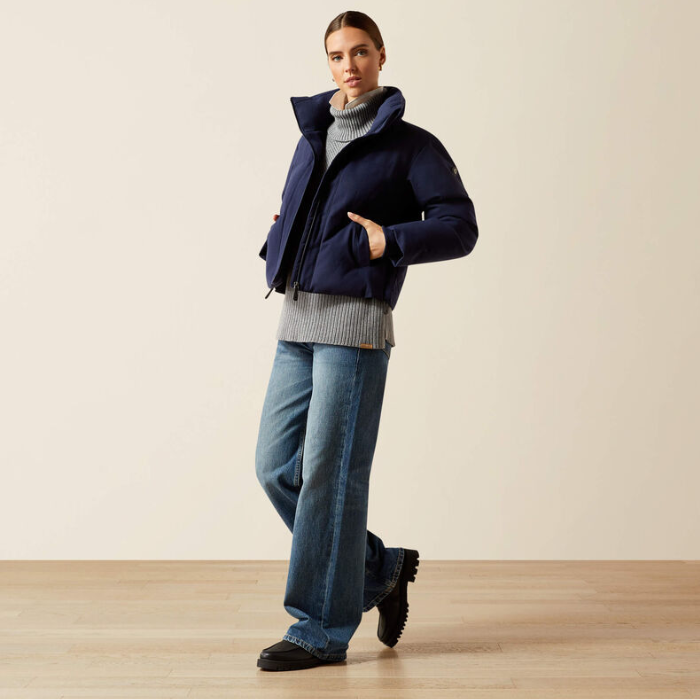 Person wearing a navy puffer jacket, grey sweater, blue jeans, and black boots on a beige background