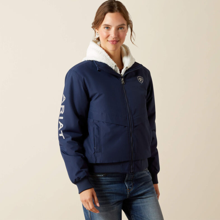 Woman wears a navy bomber jacket with Ariat branding on the sleeve on a beige background