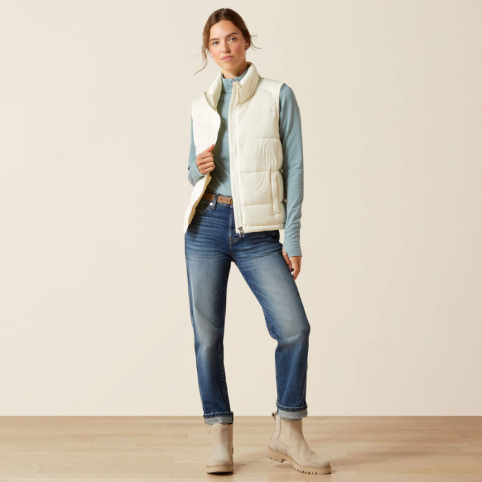 Woman wearing a light blue shirt, white puffer vest, blue jeans, and beige boots against a plain background.