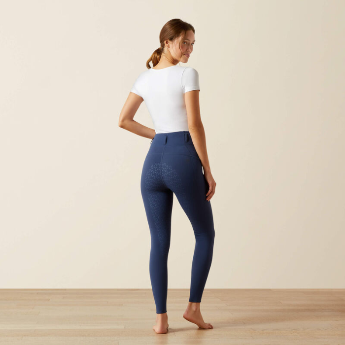 Woman wearing blue riding leggings and a white top on a plain background