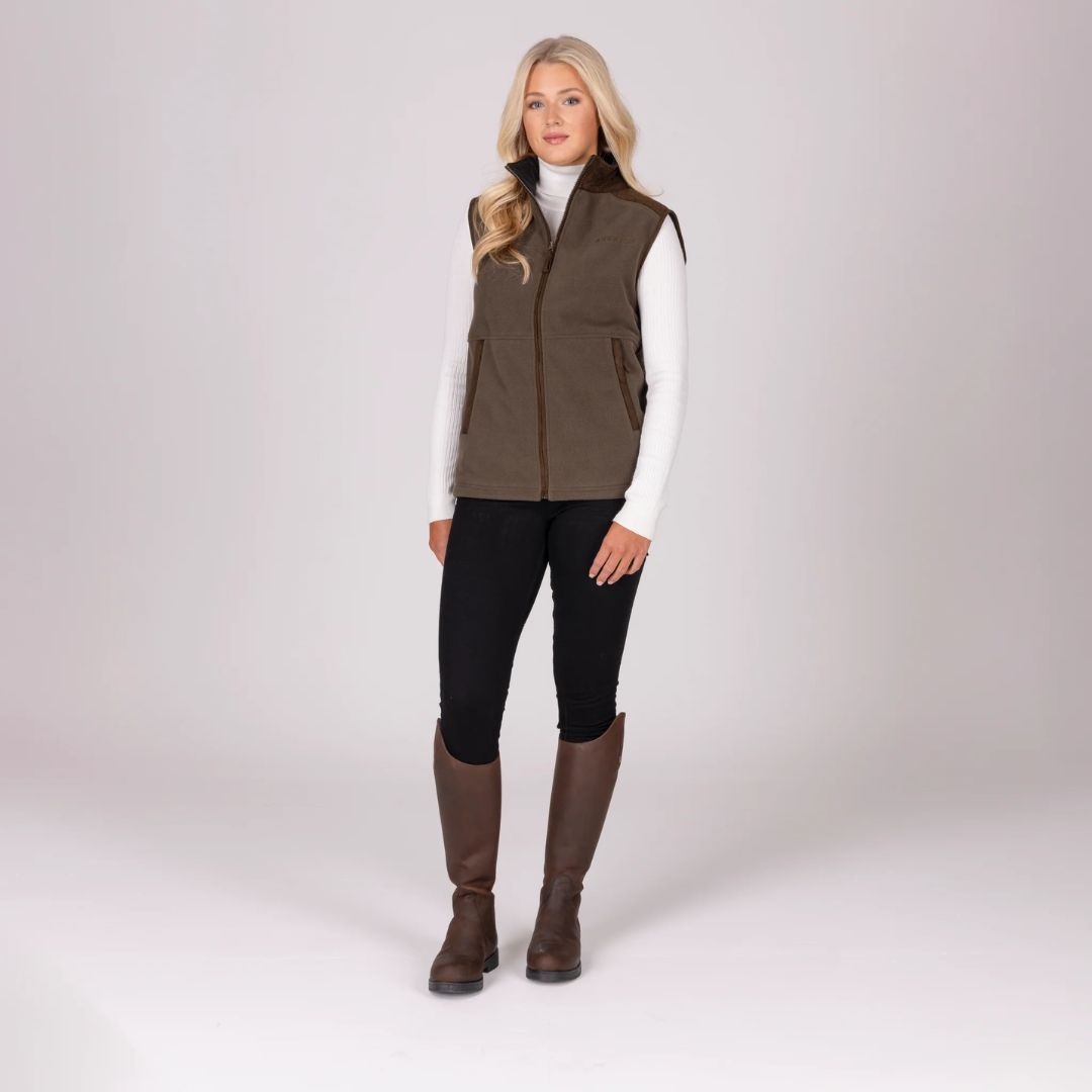 Woman wearing a brown gilet, white shirt, black leggings, and brown boots on a white background