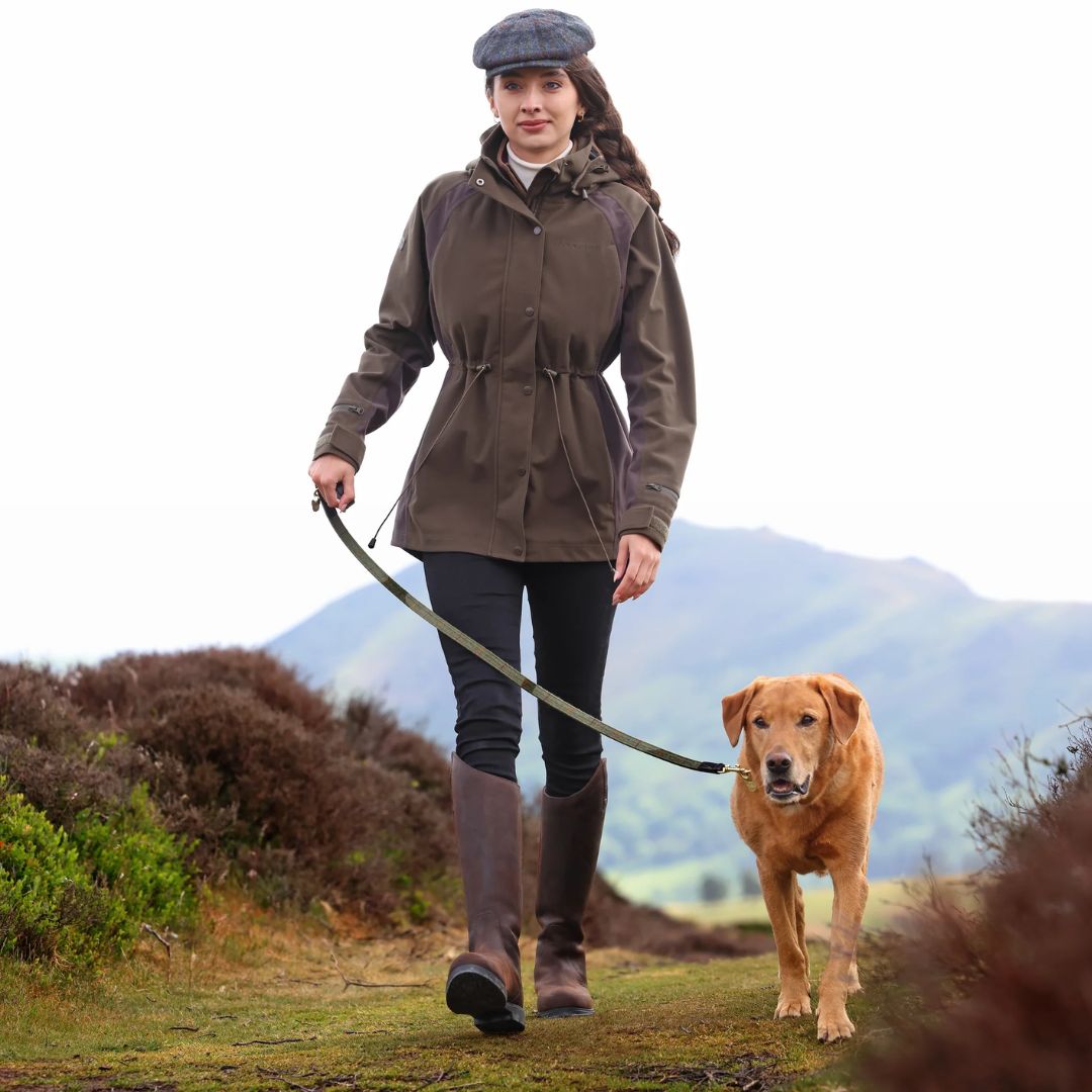 Woman in a brown jacket and flat cap walking a dog in a scenic outdoor setting