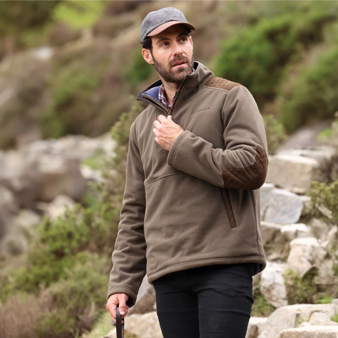 Man wearing a brown fleece jacket and cap standing in a natural setting with rocks and greenery.