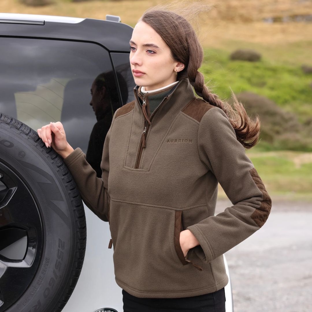 Woman in a brown fleece jacket standing next to a black vehicle in an outdoor setting
