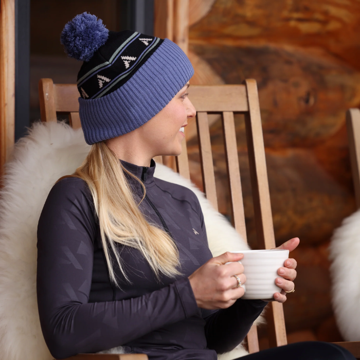 Woman wears black base layer and bobble hat whilst sat in a chair on a faux fur throw and holding a mug
