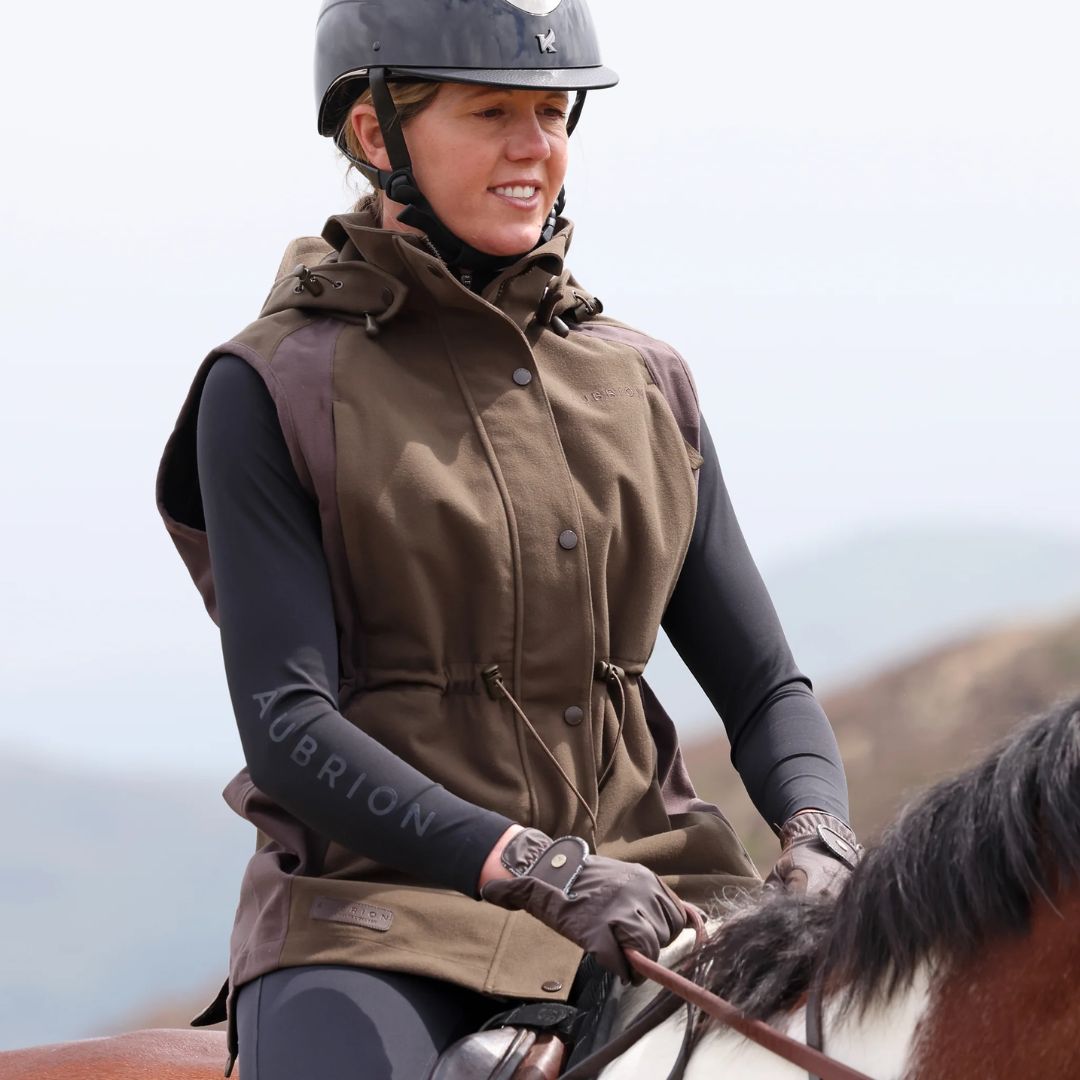 Person riding a horse wearing a brown gilet and helmet with a blurred natural background
