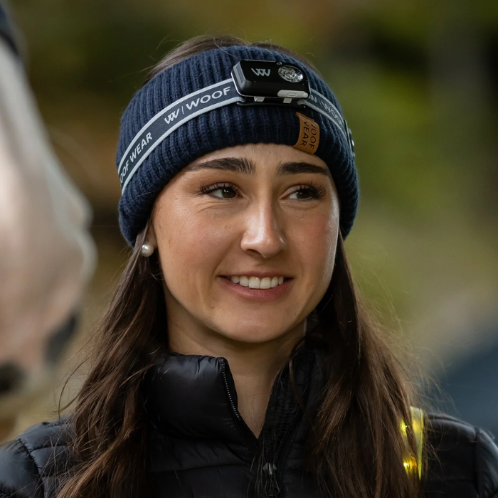 A woman wearing a navy blue ribbed knit beanie with a headlamp and a black puffer jacket, photographed outdoors with a blurred natural background.