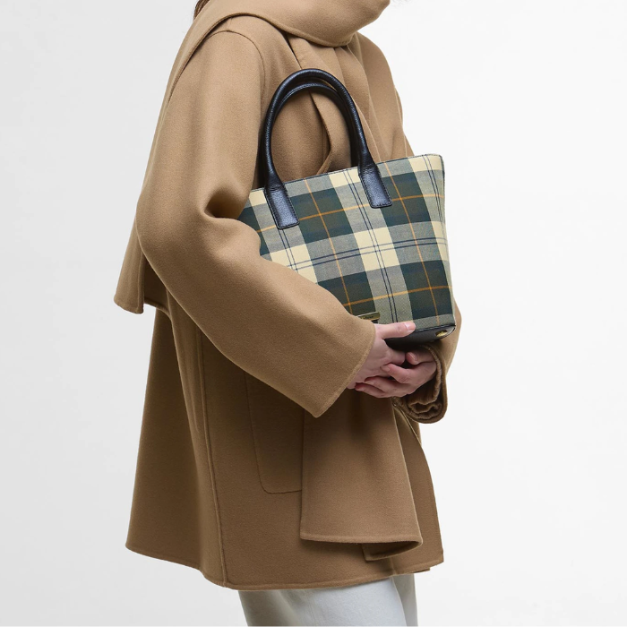 Woman clutches a tartan tote bag and wears a camel brown cape jacket on a white background