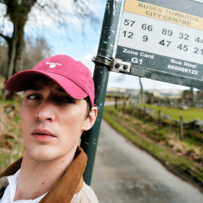 Trendy young man waits at a bus stop in the Barbour Mens Cascade Sports Cap in washed raspberry