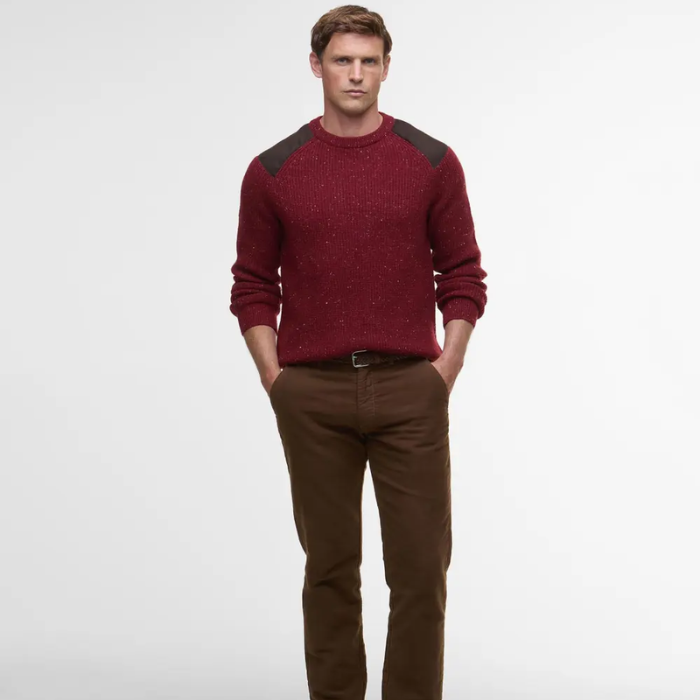 Man wearing a red Barbour jumper with shoulder detail and brown trousers on a light grey background