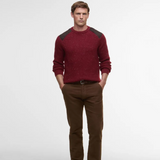 Man wearing a red Barbour jumper with shoulder detail and brown trousers on a light grey background