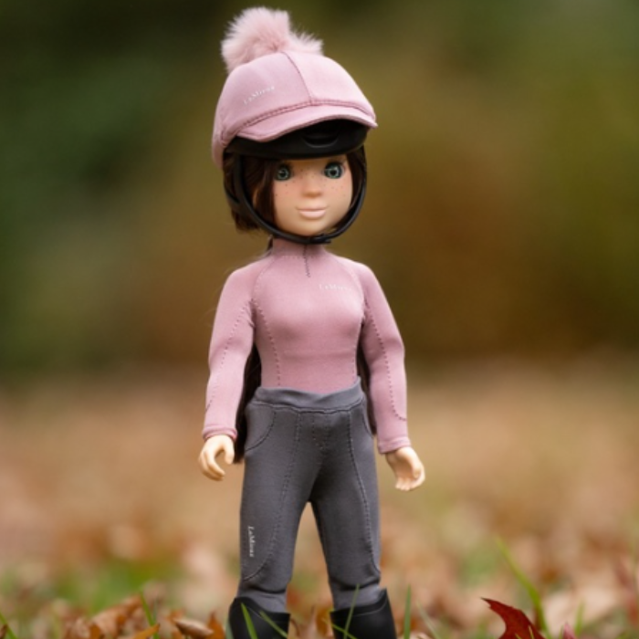 LeMieux AW26 Toy Rider Outfit Varient Blossom. Mini toy figure wearing a pink base layer, grey trousers and a matching pink hat silk stood in leaves. 