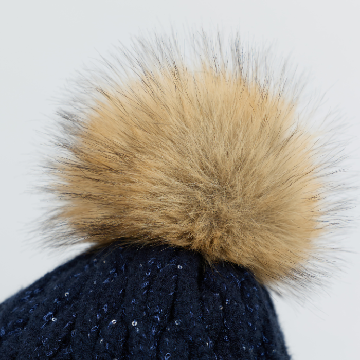 LeMieux AW26 Bernie Sequin Beanie Variant Colour Navy.
Close-up of a dark navy knit beanie topped with a large fluffy tan faux-fur pom pom, shown against a plain light gray background.