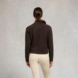 Holland Cooper Country Quarter Zip Knit Variant Colour Chocolate.
A woman viewed from behind wearing a dark brown cropped mock-neck knit sweater and cream-colored pants, standing against a textured light gray studio background.