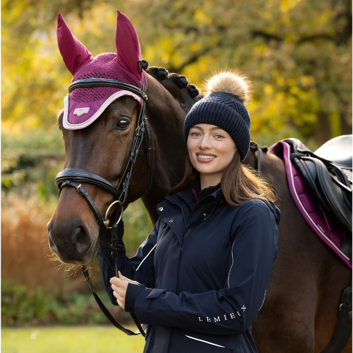 LeMieux AW26 Clara Knit Beanie Navy One Size Variant Colour Navy.
A woman in a navy equestrian jacket and navy beanie with a pom-pom stands beside a brown horse wearing a magenta fly veil and bridle in an outdoor setting with autumn trees.