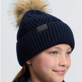 LeMieux AW26 Clara Knit Beanie Navy One Size Variant Colour Navy.
A close-up of a child wearing a navy blue ribbed knit beanie with a large tan faux-fur pom-pom. A small rectangular brand patch reading 'LeMieux' is visible on the front of the beanie.