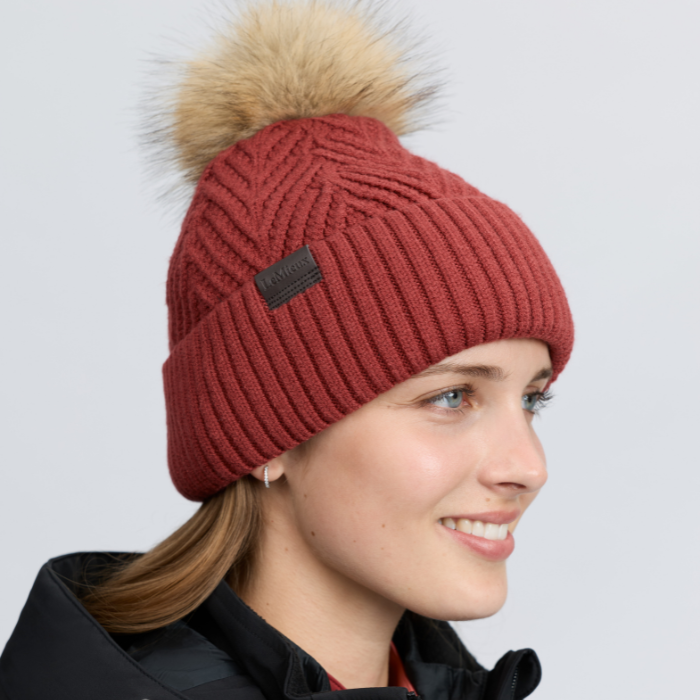 LeMieux AW26 Clara Knit Beanie Russet One Size Variant Colour Russet.
A woman wearing a ribbed knit beanie with a large tan faux-fur pom-pom. The beanie is rust red with a small black brand patch on the front, photographed against a light gray studio background.