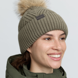 LeMieux AW26 Clara Knit Beanie Willow Green One Size Variant Colour Willow Green.
A smiling woman wearing an olive green ribbed knit beanie with a large tan faux-fur pom-pom and a small dark brand patch on the side, against a light gray background.