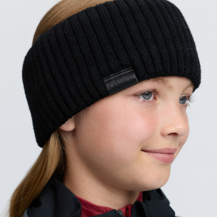 LeMieux AW26 Clara Rib Headband Variant Colouir Black.
A close-up of a child wearing a black ribbed knit headband with a small black brand tag that reads 'Bergfrost'.