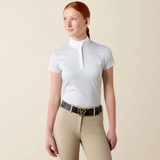 A woman modeling a white short-sleeve polo shirt with a subtle micro-check pattern, paired with beige riding pants and a black belt with a gold buckle, against a solid beige background.