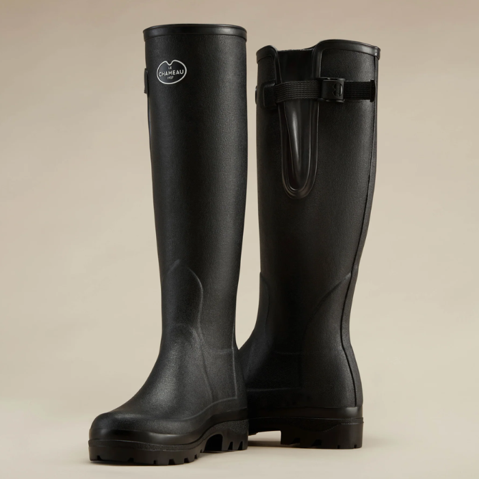 Le Chameau Ladies Vierzonord Neoprene Lined Wellies Variant Black. A pair of black rubber rain boots with a buckle strap on the back, shown against a solid beige background. One boot is standing upright to show the front profile, and the other is angled to show the back buckle strap.