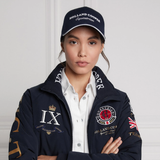 Holland Cooper HC Equestrian Cap Variant Ink Navy. Woman wearing a blue cap with 'Holland Cooper Equestrian' branding and a white trim with a matching blue coat against a white background. 