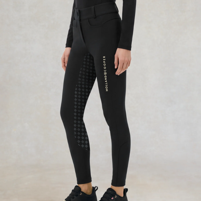 Holland Cooper Chiltern Contour Breeches Variant Black. Close up of someone wearing a pair of black breeches with Holland Cooperdown the side against a grey background. 