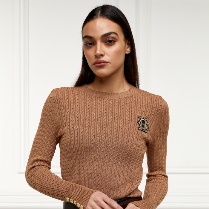 Holland Cooper Princeton Crew Neck Knit Variant Colour Dark Camel.
A woman wearing a brown cable-knit crewneck sweater with a decorative embroidered crest on the left chest, photographed against a white paneled wall.