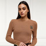 Holland Cooper Princeton Crew Neck Knit Variant Colour Dark Camel.
A woman wearing a brown cable-knit crewneck sweater with a decorative embroidered crest on the left chest, photographed against a white paneled wall.