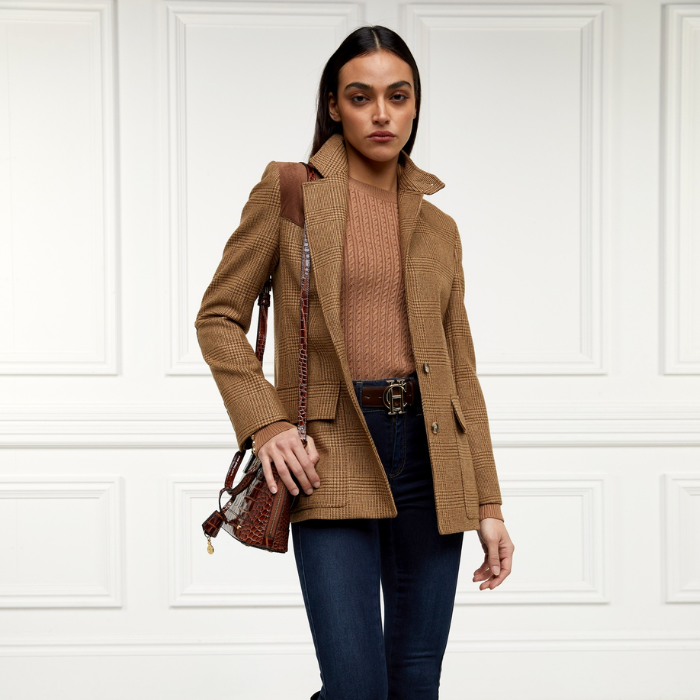 Holland Cooper Princeton Crew Neck Knit Variant Colour Dark Camel.
A woman wearing a brown cable-knit crewneck sweater, a brown jacket and over shouler handbag and dark blue jeans.  Photographed against a white paneled wall.