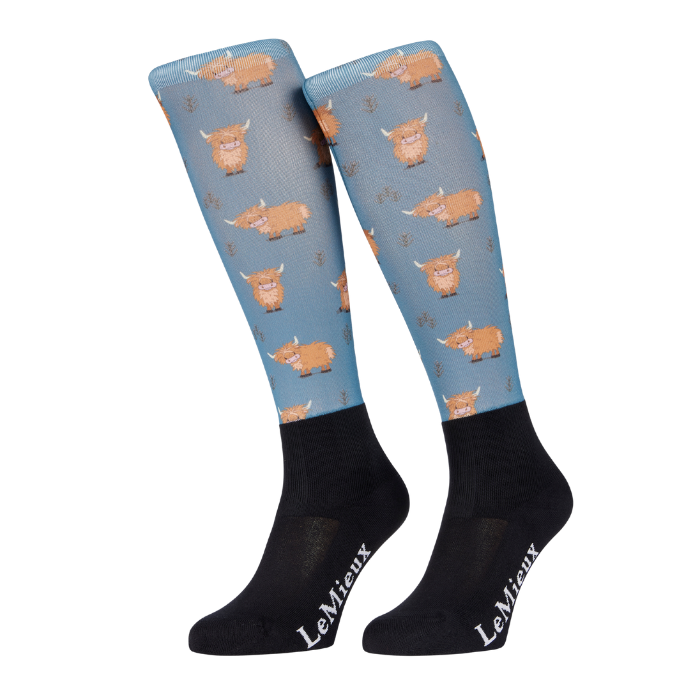 LeMieux AW26 Footsie Socks Highland Cow Vairant Colour Aloe.
A pair of LeMieux knee-high socks featuring a light blue upper leg section with a repeating pattern of brown highland cows, and solid black lower legs with white LeMieux branding.