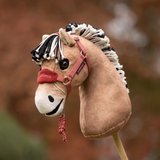 LeMieux AW26 Hobby Horse Vogue Headcollar & Leadrope Variant Colour Spiced Earth. 
A plush toy horse head mounted on a stick, with a white body, gray faux-fur mane, and Spiced earth colour halter, photographed outdoors against a blurred garden background.